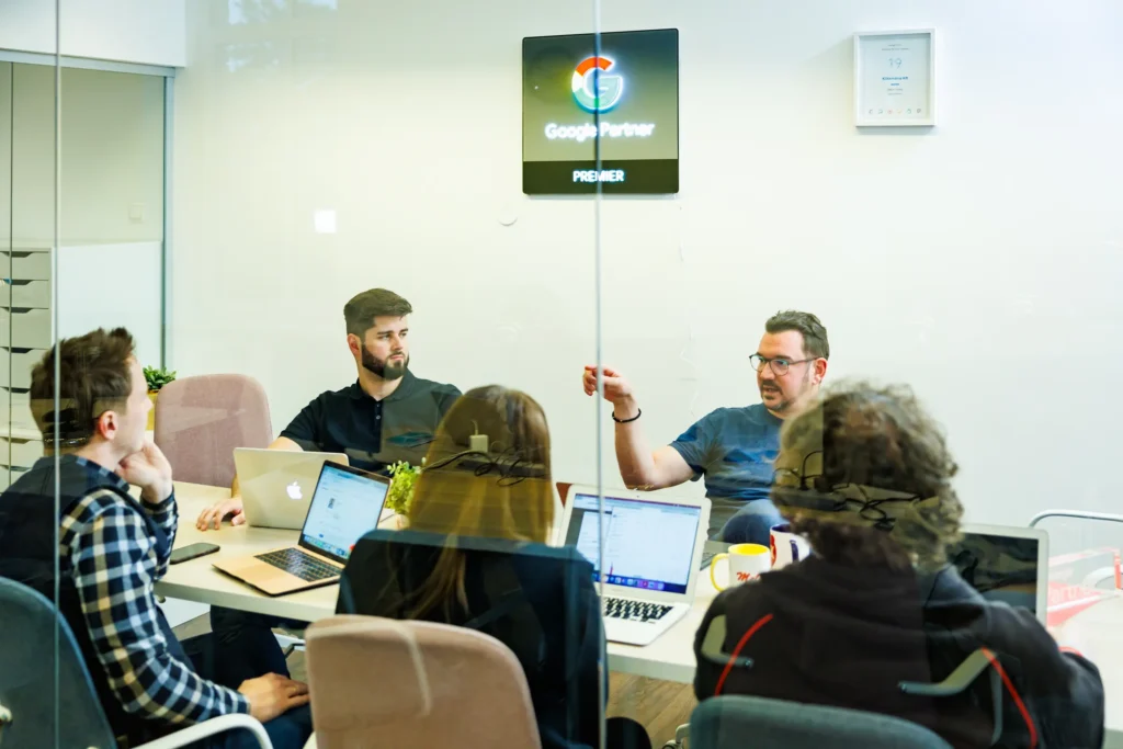 Egy csoport ember ül egy tárgyalóasztal körül laptoppal. Egy ember beszél, míg mások hallgatnak. A falon egy „Google Partner Premier” felirat látható. A szoba üvegfalú, az asztalon cserepes növények vannak.