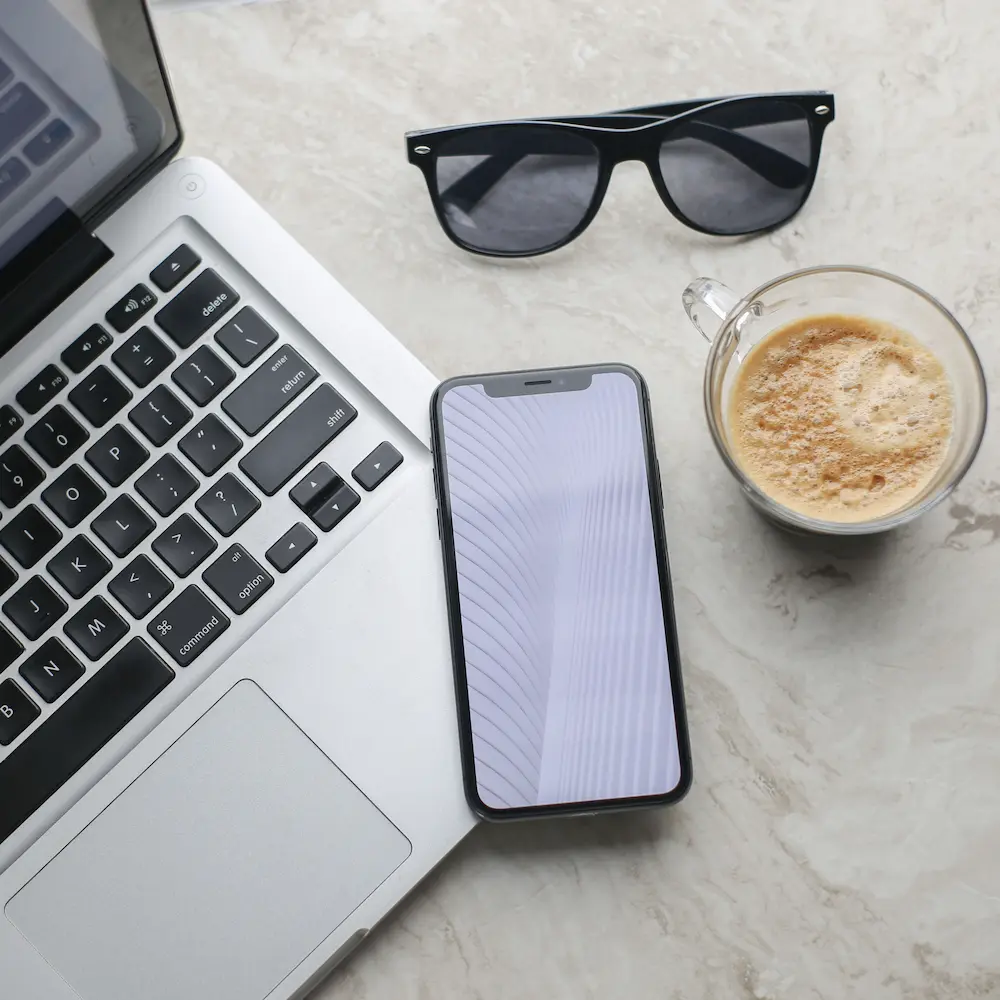 Egy laptop, amelyen látható a billentyűzet, egy okostelefon mellett halvány vonalak láthatók, ami a Google Ads-statisztikákra utal. A közelben fekete napszemüveg és habos kávéval töltött üvegbögre pihen a világos márványfelületen, készen áll a következő kampánystratégia ötletelésre.