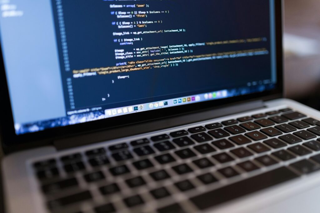 Egy laptop programozási kódot jelenít meg a képernyőjén, ami egy WordPress környezetre emlékeztet. A billentyűzet részben látható, a képernyőre fókuszálva, a kódolási munkaterületre jellemző többszínű szövegsorok láthatók. A háttér kissé elmosódott, kiemelve a Core Web Vitals mutatót működés közben.