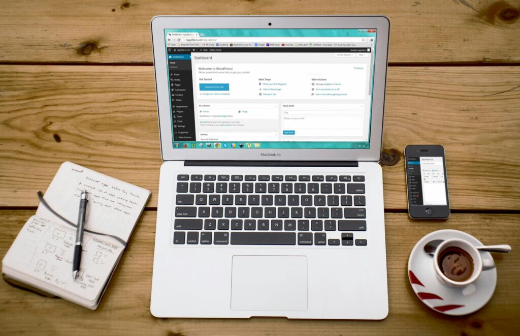 Egy WordPress irányítópultot bemutató laptop egy fából készült íróasztalon, egy oldalsó jegyzettömbön lévő vázlatok, egy okostelefon és egy gőzölgő kávé mellett ül. Ez a dinamikus munkaterület az aktív webhelykezelésre vagy tartalomkészítésre utal, szem előtt tartva a Core Web Vitals-t.